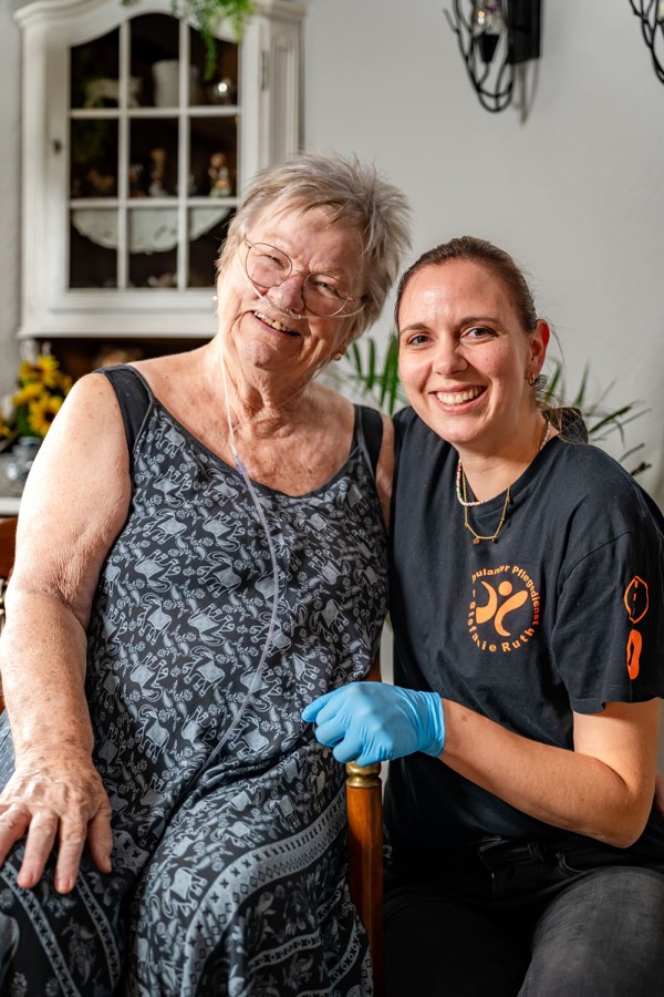 Eine Kundin und eine Pflegekraft in dunklem Dienstshirt sitzen Wange an Wange und lächeln in die Kamera