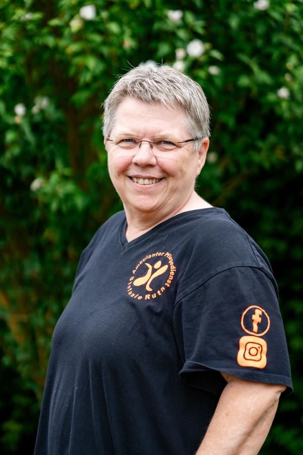 Eine Pflegekraft mit kurzen grauen Haaren in dunklem Dienstshirt lächelt im Garten