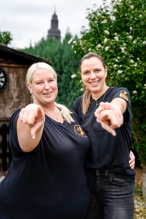 Zwei Kolleginnen in dunklen Dienstshirts zeigen lachend auf den Betrachter