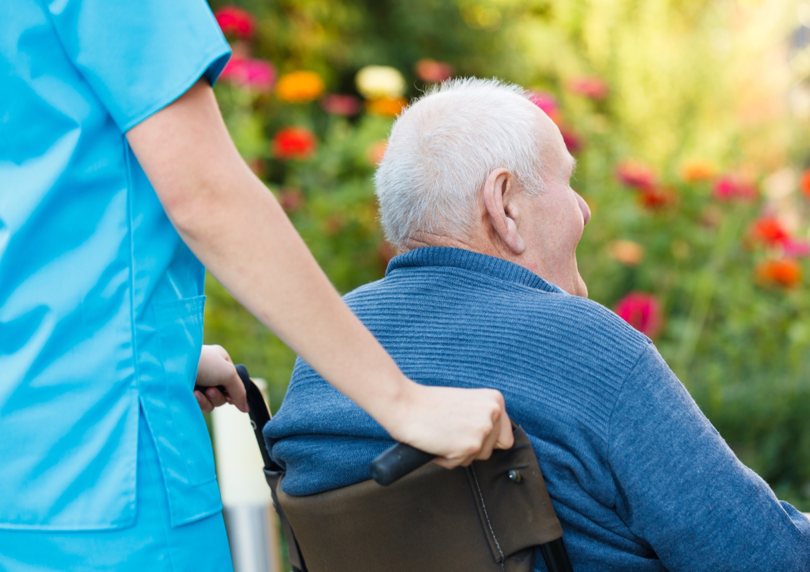 Eine Pflegekraft begleitet einen älteren Mann im Rollstuhl durch einen Garten