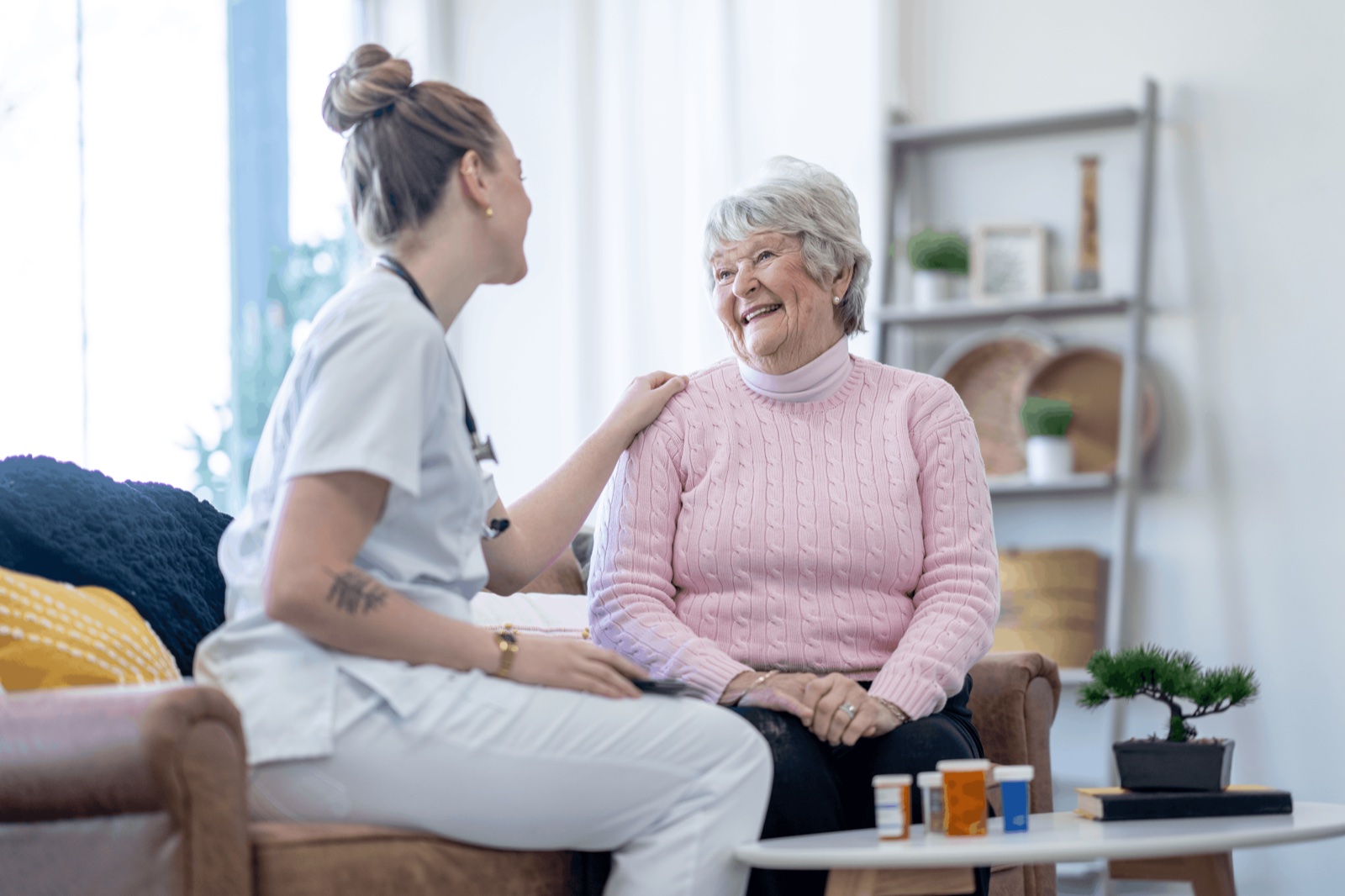 Eine Pflegerin sitzt zugewandt neben einer lachenden Seniorin im Wohnzimmer
