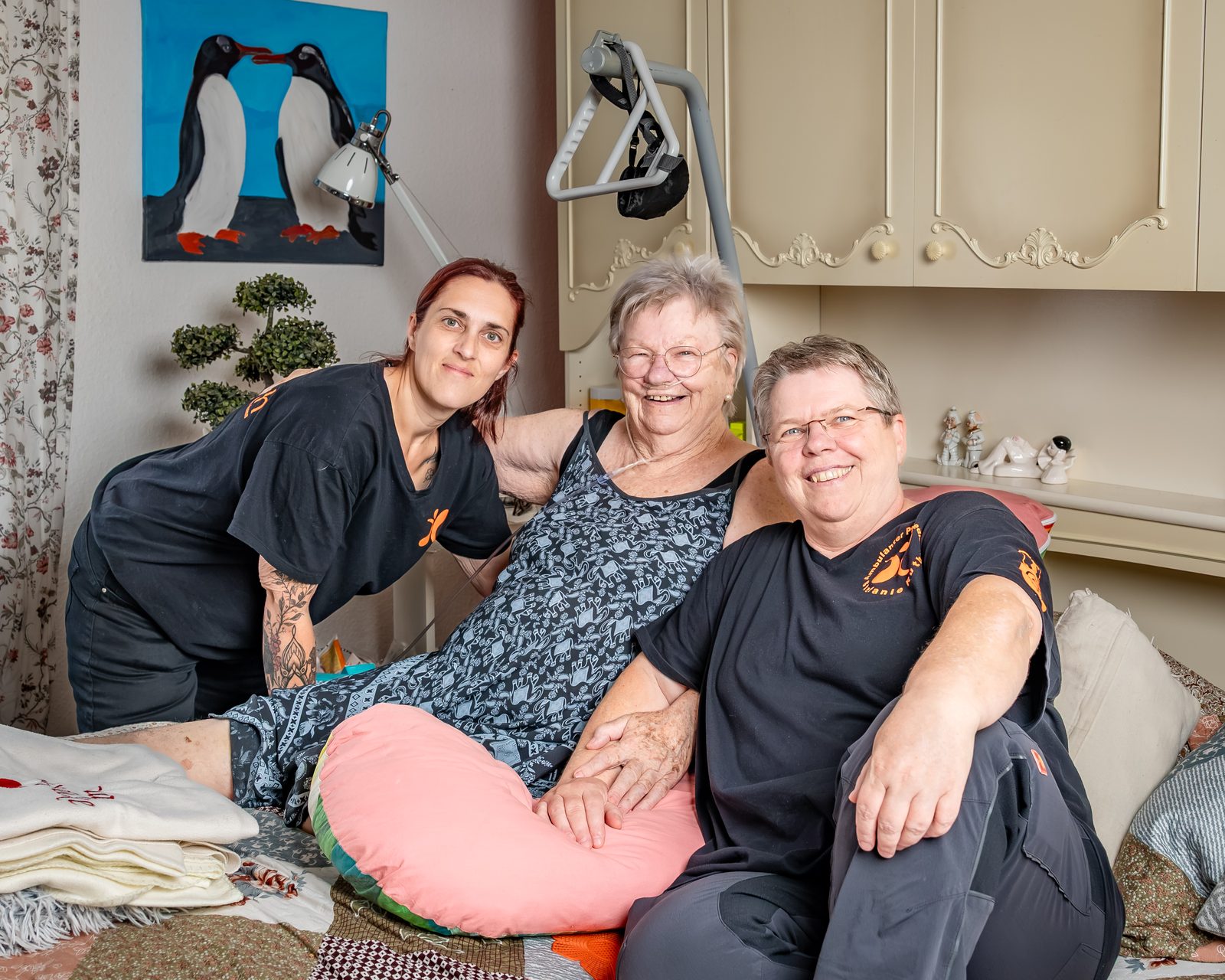 Inhaberin Stefanie Ruth und eine Kollegin sitzen mit einer Kundin lächelnd auf dem Bett in deren Zuhause